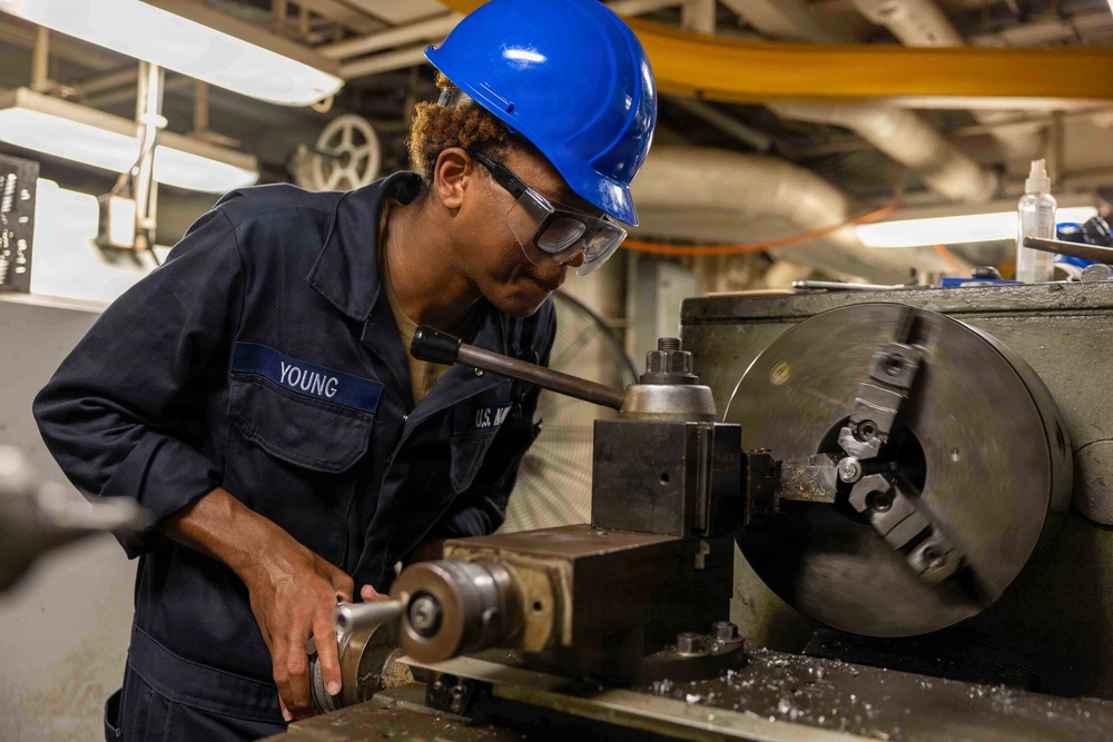 A U.S. Navy service member with the last name 'Young' is operating a metal lathe in what appears to be a shipboard machine shop. The individual is wearing standard Navy coveralls, a blue hard hat, and safety goggles while working with precision machinery. The setting suggests an industrial workspace aboard a naval vessel.