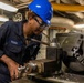 A U.S. Navy service member with the last name 'Young' is operating a metal lathe in what appears to be a shipboard machine shop. The individual is wearing standard Navy coveralls, a blue hard hat, and safety goggles while working with precision machinery. The setting suggests an industrial workspace aboard a naval vessel.