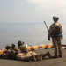 Three military personnel in desert camouflage uniforms are positioned on what appears to be the deck of a naval vessel overlooking open water. Two soldiers are in a prone firing position while a third stands upright facing the water, all equipped with tactical gear and weapons. The scene suggests a live-fire exercise or security drill conducted from a ship's deck.