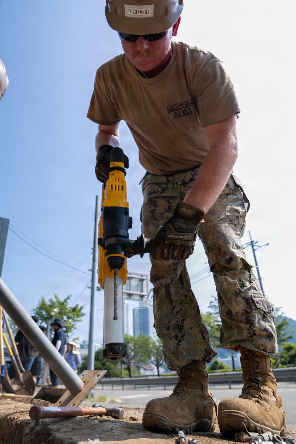 A military servicemember identified as 'Richard' wearing a tan hard hat, sunglasses, camouflage pants, and an NMCB (Naval Mobile Construction Battalion) t-shirt operates a yellow DeWalt rotary hammer drill at an outdoor construction site. He is wearing Mechanix M-PACT gloves and tan military boots while drilling into a surface. Other workers are visible in the background along with urban infrastructure including buildings, trees, and utility poles.