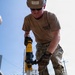 A military servicemember identified as 'Richard' wearing a tan hard hat, sunglasses, camouflage pants, and an NMCB (Naval Mobile Construction Battalion) t-shirt operates a yellow DeWalt rotary hammer drill at an outdoor construction site. He is wearing Mechanix M-PACT gloves and tan military boots while drilling into a surface. Other workers are visible in the background along with urban infrastructure including buildings, trees, and utility poles.