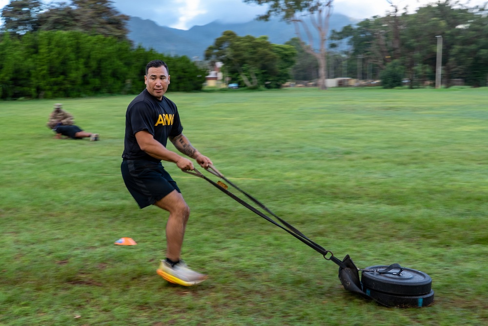 A man wearing a black 'ARMY' t-shirt and shorts is pulling a weighted sled across a grass field using resistance straps, performing a strength and conditioning exercise. The setting appears to be a military physical training session on an open field with mountains and trees visible in the background. Another person can be seen lying on the ground in the background, likely performing a separate exercise.