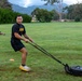 A man wearing a black 'ARMY' t-shirt and shorts is pulling a weighted sled across a grass field using resistance straps, performing a strength and conditioning exercise. The setting appears to be a military physical training session on an open field with mountains and trees visible in the background. Another person can be seen lying on the ground in the background, likely performing a separate exercise.
