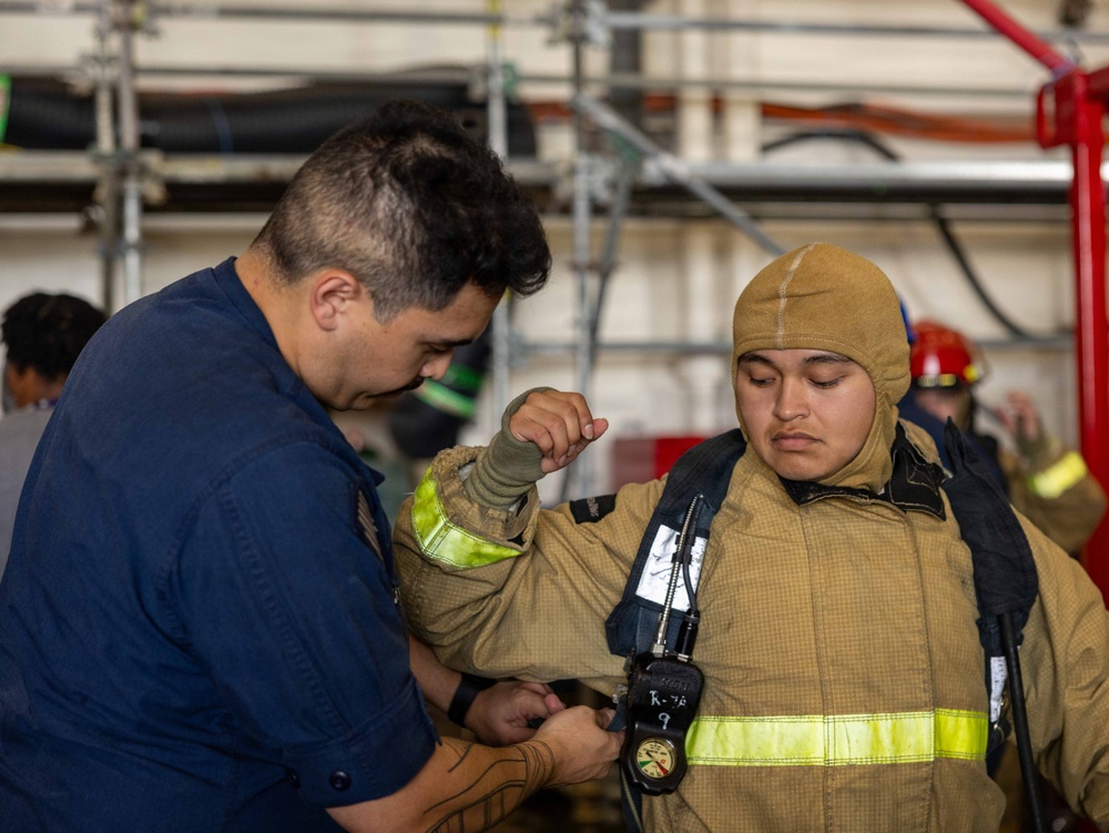 Two individuals are shown in what appears to be a firefighting training environment. One person wearing a navy blue uniform is assisting another person who is dressed in full firefighting gear including a tan/gold protective suit, hood, and SCBA (Self-Contained Breathing Apparatus) equipment. The setting appears to be an industrial or shipboard training facility with scaffolding and hoses visible in the background.