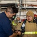 Two individuals are shown in what appears to be a firefighting training environment. One person wearing a navy blue uniform is assisting another person who is dressed in full firefighting gear including a tan/gold protective suit, hood, and SCBA (Self-Contained Breathing Apparatus) equipment. The setting appears to be an industrial or shipboard training facility with scaffolding and hoses visible in the background.