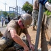 A military service member wearing a tan helmet and camouflage uniform is kneeling on the ground securing a metal post/pole to a base plate using fasteners. Additional personnel in similar uniforms are visible in the background along a wall. The work appears to be a construction or infrastructure installation project in an outdoor urban setting.