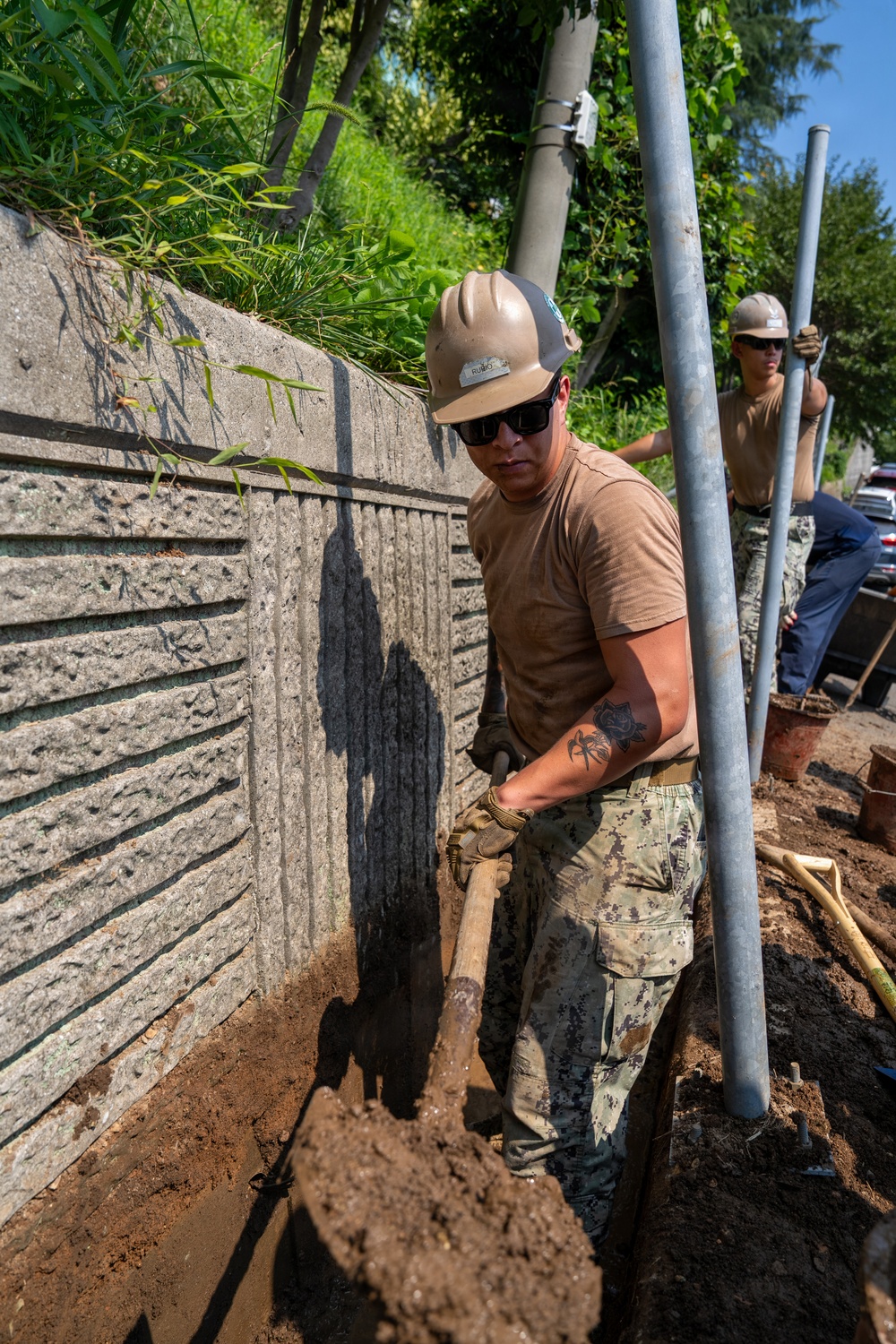 Two workers in military-style camouflage uniforms and tan hard hats are performing excavation work along a concrete retaining wall. The foreground worker is actively shoveling dirt in a narrow trench while a second worker in the background handles metal poles or pipes. The setting appears to be an outdoor construction or utility installation site with lush vegetation visible above the wall.