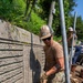 Two workers in military-style camouflage uniforms and tan hard hats are performing excavation work along a concrete retaining wall. The foreground worker is actively shoveling dirt in a narrow trench while a second worker in the background handles metal poles or pipes. The setting appears to be an outdoor construction or utility installation site with lush vegetation visible above the wall.