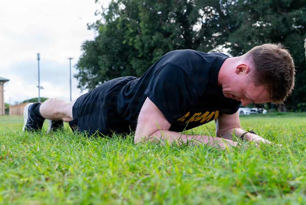 A male individual wearing a black U.S. Army physical training shirt is performing push-ups on a grass field outdoors. He is in the lowered position of the exercise, with his body close to the ground. The setting appears to be a military installation or training area based on the attire and background infrastructure.
