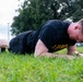 A male individual wearing a black U.S. Army physical training shirt is performing push-ups on a grass field outdoors. He is in the lowered position of the exercise, with his body close to the ground. The setting appears to be a military installation or training area based on the attire and background infrastructure.