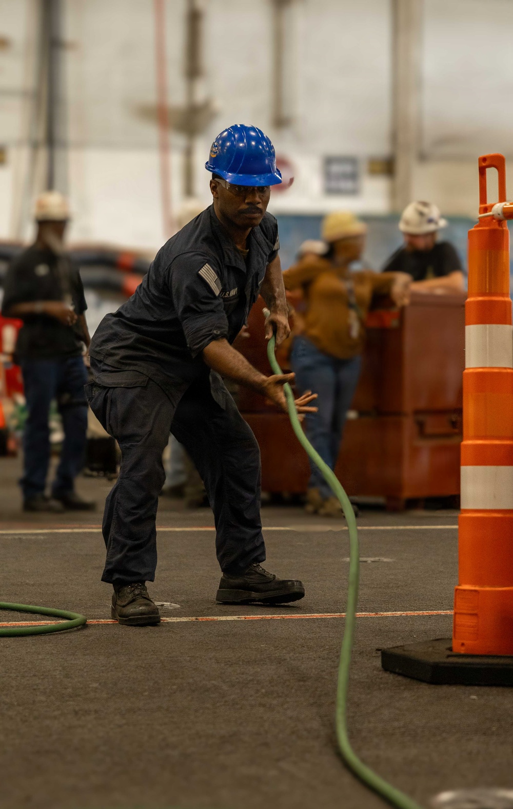 A U.S. Navy sailor wearing dark blue coveralls and a blue hard hat is pulling a green hose or rope in what appears to be an indoor industrial or hangar bay environment. The scene suggests a damage control or firefighting drill, with orange traffic cones marking a course area. Several other personnel in the background are also engaged in related activities.
