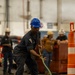 A U.S. Navy sailor wearing dark blue coveralls and a blue hard hat is pulling a green hose or rope in what appears to be an indoor industrial or hangar bay environment. The scene suggests a damage control or firefighting drill, with orange traffic cones marking a course area. Several other personnel in the background are also engaged in related activities.