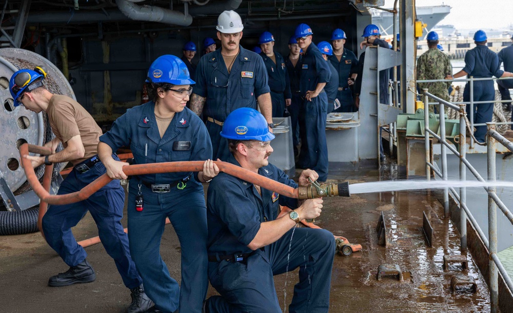 Naval personnel in dark blue uniforms and blue hard hats are conducting a firefighting drill or damage control exercise aboard a military vessel. Two sailors in the foreground are actively operating a fire hose connected to a brass nozzle, with water visibly spraying onto the wet deck. A group of observers in similar uniforms watches from the background, suggesting this is a training evaluation or demonstration.