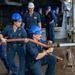 Naval personnel in dark blue uniforms and blue hard hats are conducting a firefighting drill or damage control exercise aboard a military vessel. Two sailors in the foreground are actively operating a fire hose connected to a brass nozzle, with water visibly spraying onto the wet deck. A group of observers in similar uniforms watches from the background, suggesting this is a training evaluation or demonstration.