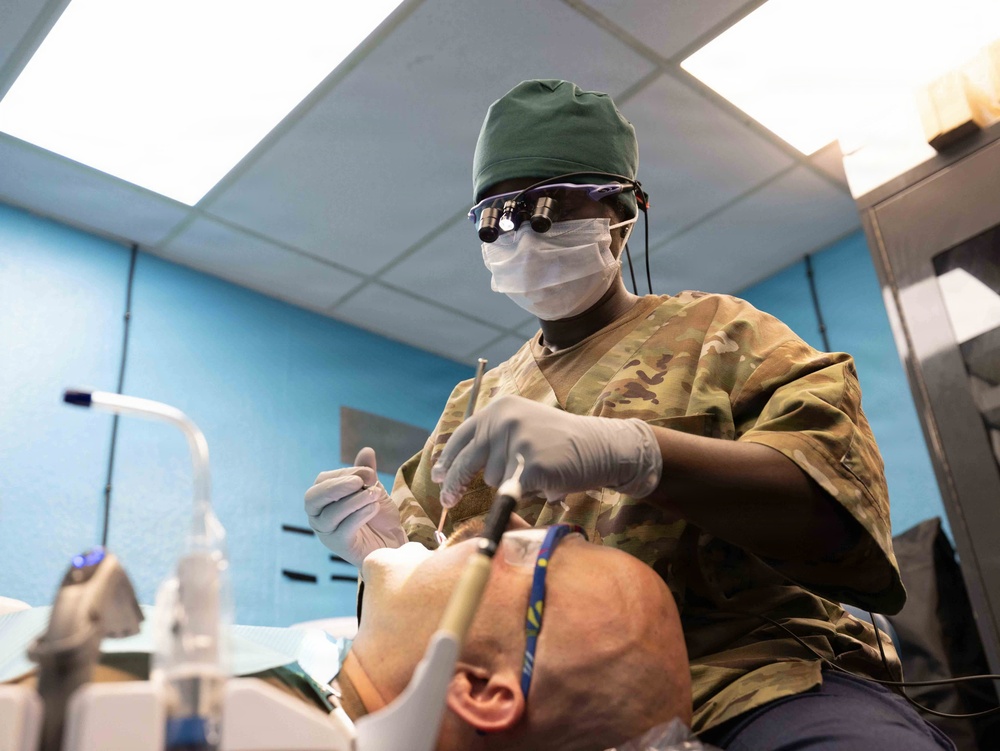 A military dental professional wearing OCP (Operational Camouflage Pattern) uniform is performing a dental procedure on a reclined patient. The practitioner is equipped with surgical loupes, a green scrub cap, face mask, and latex gloves, working in what appears to be a military dental clinic. The low-angle shot provides a patient's-eye perspective of the procedure.