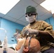 A military dental professional wearing OCP (Operational Camouflage Pattern) uniform is performing a dental procedure on a reclined patient. The practitioner is equipped with surgical loupes, a green scrub cap, face mask, and latex gloves, working in what appears to be a military dental clinic. The low-angle shot provides a patient's-eye perspective of the procedure.