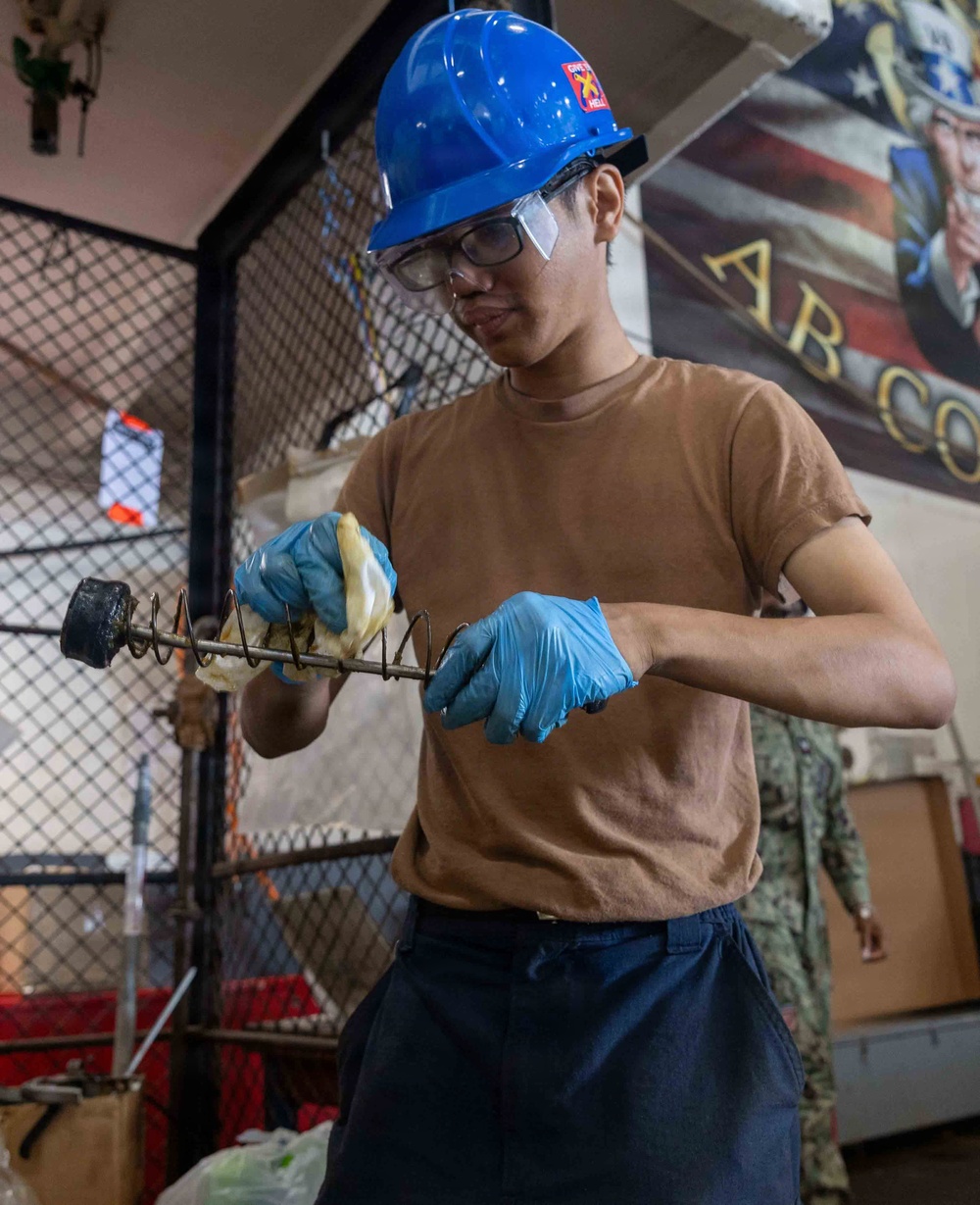 A young person wearing a blue hard hat, safety glasses, and blue nitrile gloves is cleaning or maintaining a mechanical component, likely a spring or coil assembly, using a white cloth or rag. They are dressed in a tan t-shirt and navy blue pants, suggesting a military or industrial work environment. In the background, a partial U.S. military recruitment or unit poster is visible along with chain-link fencing and equipment.