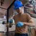 A young person wearing a blue hard hat, safety glasses, and blue nitrile gloves is cleaning or maintaining a mechanical component, likely a spring or coil assembly, using a white cloth or rag. They are dressed in a tan t-shirt and navy blue pants, suggesting a military or industrial work environment. In the background, a partial U.S. military recruitment or unit poster is visible along with chain-link fencing and equipment.