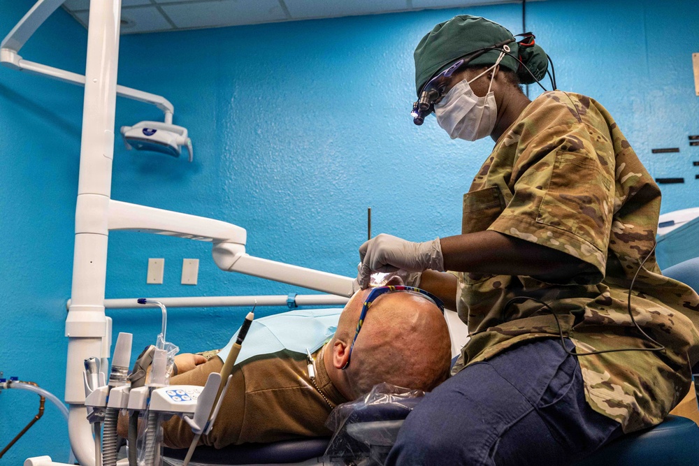 A military dental professional in U.S. Army OCP (Operational Camouflage Pattern) uniform is performing a dental procedure on a reclined patient. The practitioner is wearing full personal protective equipment including a surgical mask, green scrub cap, latex gloves, and magnification loupes with an attached light. The setting appears to be a military or field dental clinic with characteristic blue walls and standard dental equipment.