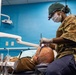 A military dental professional in U.S. Army OCP (Operational Camouflage Pattern) uniform is performing a dental procedure on a reclined patient. The practitioner is wearing full personal protective equipment including a surgical mask, green scrub cap, latex gloves, and magnification loupes with an attached light. The setting appears to be a military or field dental clinic with characteristic blue walls and standard dental equipment.