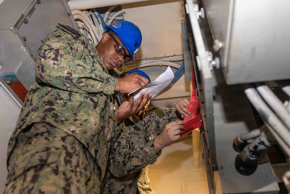 Two U.S. Navy personnel in digital camouflage uniforms are working in a confined mechanical or equipment space aboard what appears to be a naval vessel. One sailor wearing a blue hard hat and safety glasses is reviewing a document while the other, identified by a name tape reading 'WATSON', is handling a red DANGER lockout/tagout tag on equipment. They appear to be conducting a safety lockout/tagout procedure on shipboard machinery.