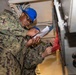 Two U.S. Navy personnel in digital camouflage uniforms are working in a confined mechanical or equipment space aboard what appears to be a naval vessel. One sailor wearing a blue hard hat and safety glasses is reviewing a document while the other, identified by a name tape reading 'WATSON', is handling a red DANGER lockout/tagout tag on equipment. They appear to be conducting a safety lockout/tagout procedure on shipboard machinery.