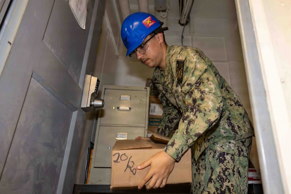 A U.S. Navy sailor wearing NWU Type III camouflage uniform and a blue hard hat is handling a cardboard box marked '20/20' in what appears to be a confined shipboard compartment or stairwell. The sailor is wearing safety glasses and appears to be moving or organizing supplies. The setting suggests a logistics or supply operation aboard a naval vessel.