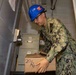 A U.S. Navy sailor wearing NWU Type III camouflage uniform and a blue hard hat is handling a cardboard box marked '20/20' in what appears to be a confined shipboard compartment or stairwell. The sailor is wearing safety glasses and appears to be moving or organizing supplies. The setting suggests a logistics or supply operation aboard a naval vessel.