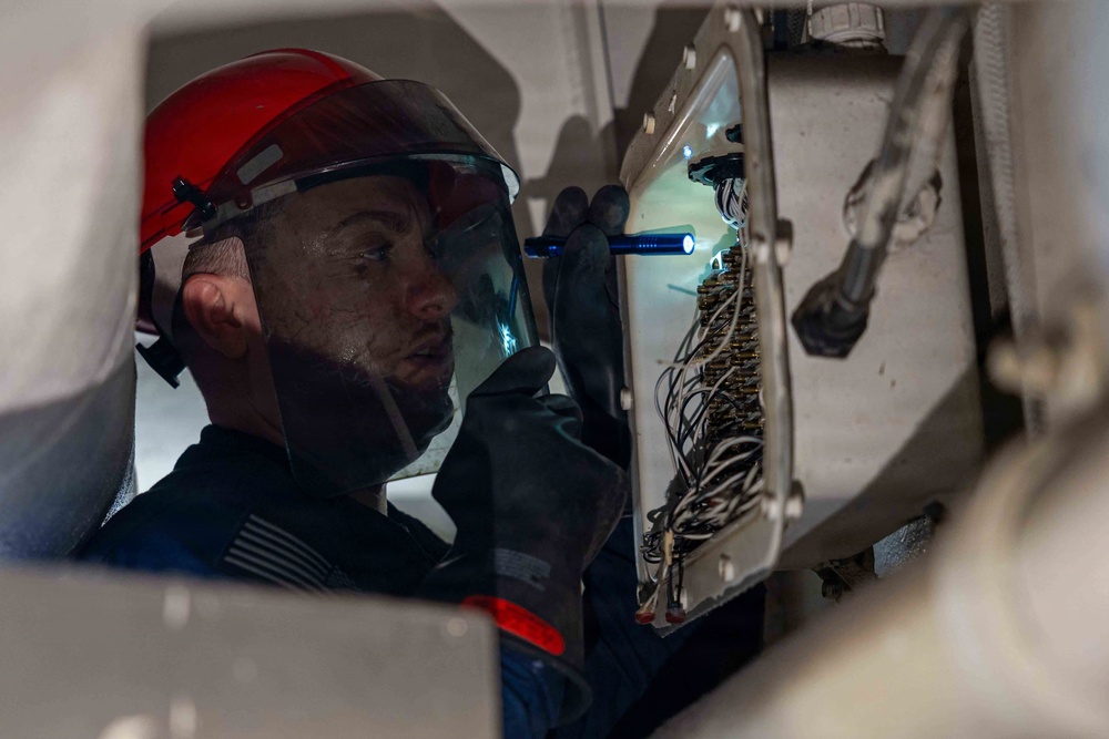 A technician wearing a red hard hat and face shield is inspecting an open electrical or wiring panel using a small flashlight. The worker appears to be in a confined or enclosed space, carefully examining a dense array of wires and terminals. A U.S. flag patch is visible on the sleeve, suggesting military or government affiliation.
