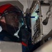 A technician wearing a red hard hat and face shield is inspecting an open electrical or wiring panel using a small flashlight. The worker appears to be in a confined or enclosed space, carefully examining a dense array of wires and terminals. A U.S. flag patch is visible on the sleeve, suggesting military or government affiliation.