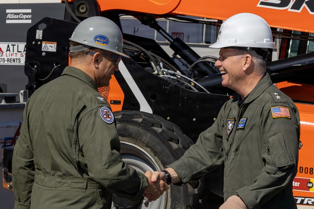 Two individuals wearing olive/sage green flight suits and hard hats are shaking hands in front of orange construction equipment. Both appear to be military personnel based on their flight suits, unit patches, and rank insignia. The person on the right wears a white hard hat and displays a two-star rank insignia along with a US flag patch, suggesting a senior military officer.