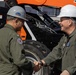Two individuals wearing olive/sage green flight suits and hard hats are shaking hands in front of orange construction equipment. Both appear to be military personnel based on their flight suits, unit patches, and rank insignia. The person on the right wears a white hard hat and displays a two-star rank insignia along with a US flag patch, suggesting a senior military officer.