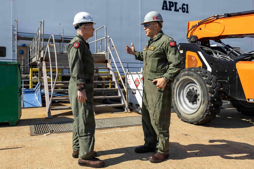 Two military personnel in olive green flight suits and white hard hats are engaged in conversation on what appears to be a naval facility or pier. They are standing near a vessel or structure marked 'APL-61' with an orange telehandler/forklift visible in the background. Both individuals wear patches on their uniforms and appear to be conducting an inspection or briefing.