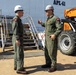 Two military personnel in olive green flight suits and white hard hats are engaged in conversation on what appears to be a naval facility or pier. They are standing near a vessel or structure marked 'APL-61' with an orange telehandler/forklift visible in the background. Both individuals wear patches on their uniforms and appear to be conducting an inspection or briefing.