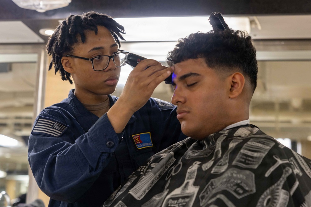 A person in a U.S. military uniform is cutting the hair of a seated client in what appears to be a barbershop setting. The barber is wearing a Navy blue uniform with a U.S. flag patch on the sleeve and a unit insignia patch on the chest. The client is draped in a barber's cape and has his eyes closed while receiving a haircut with electric clippers.