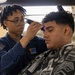 A person in a U.S. military uniform is cutting the hair of a seated client in what appears to be a barbershop setting. The barber is wearing a Navy blue uniform with a U.S. flag patch on the sleeve and a unit insignia patch on the chest. The client is draped in a barber's cape and has his eyes closed while receiving a haircut with electric clippers.