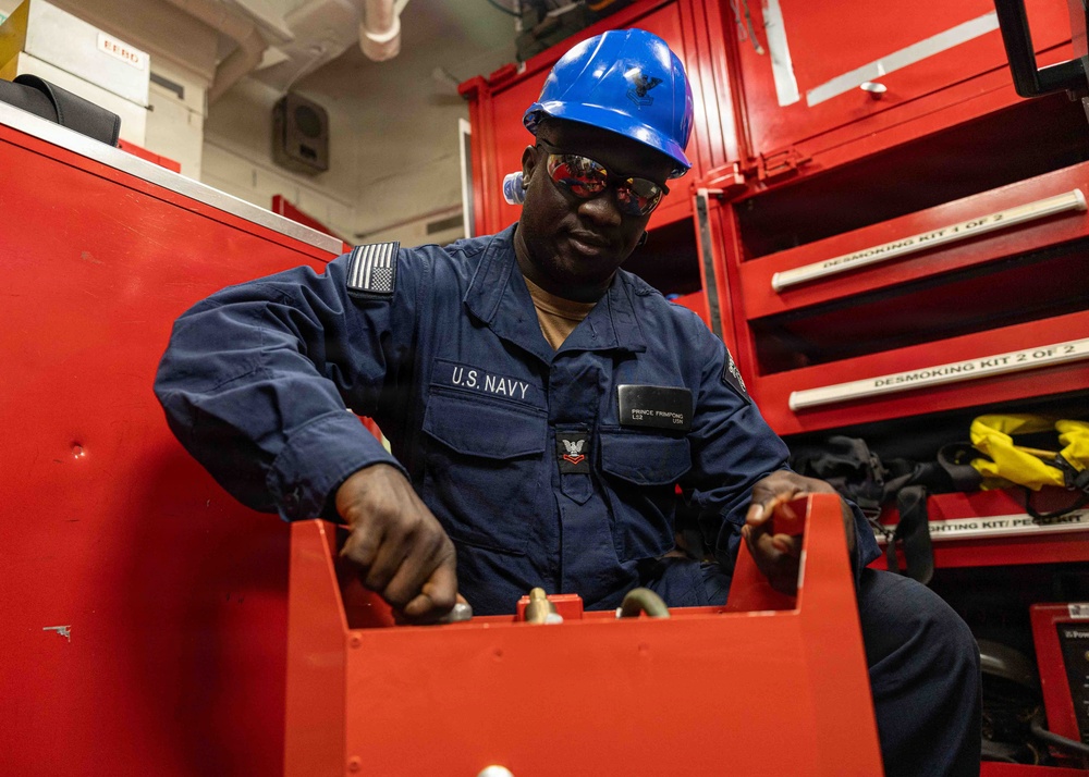 A U.S. Navy sailor wearing a blue coverall uniform and blue hard hat is working with red equipment in what appears to be a shipboard workspace. The sailor's name tag reads 'PRINCE FRIMPONG' with a rate/rank insignia visible on the chest. The background shows red tool storage cabinets labeled with 'DESMOKING KIT 1 OF 2' and 'DESMOKING KIT 2 OF 2'.