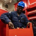A U.S. Navy sailor wearing a blue coverall uniform and blue hard hat is working with red equipment in what appears to be a shipboard workspace. The sailor's name tag reads 'PRINCE FRIMPONG' with a rate/rank insignia visible on the chest. The background shows red tool storage cabinets labeled with 'DESMOKING KIT 1 OF 2' and 'DESMOKING KIT 2 OF 2'.