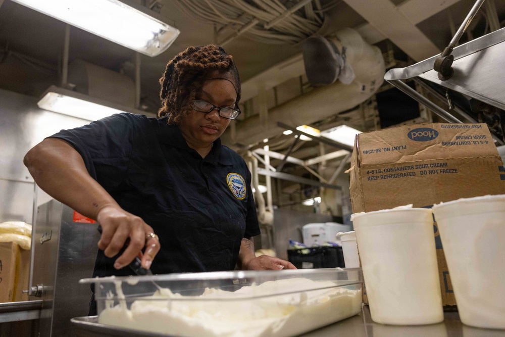 A woman wearing a dark navy uniform with an official patch is working in what appears to be a military ship's galley kitchen. She is handling food preparation, working with what appears to be sour cream or a similar dairy product in a metal tray. The industrial kitchen environment features exposed piping, fluorescent lighting, and stainless steel equipment.