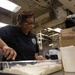 A woman wearing a dark navy uniform with an official patch is working in what appears to be a military ship's galley kitchen. She is handling food preparation, working with what appears to be sour cream or a similar dairy product in a metal tray. The industrial kitchen environment features exposed piping, fluorescent lighting, and stainless steel equipment.