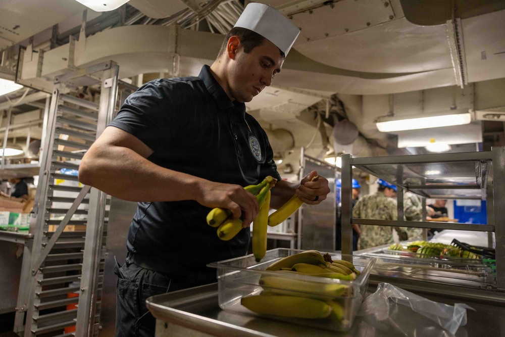 A food service worker wearing a white paper chef's hat and black polo shirt is peeling bananas in what appears to be a military ship's galley (kitchen). Several bananas are arranged in a stainless steel serving tray in front of him. In the background, personnel in Navy working uniforms are visible, along with industrial kitchen equipment and storage racks.