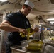 A food service worker wearing a white paper chef's hat and black polo shirt is peeling bananas in what appears to be a military ship's galley (kitchen). Several bananas are arranged in a stainless steel serving tray in front of him. In the background, personnel in Navy working uniforms are visible, along with industrial kitchen equipment and storage racks.