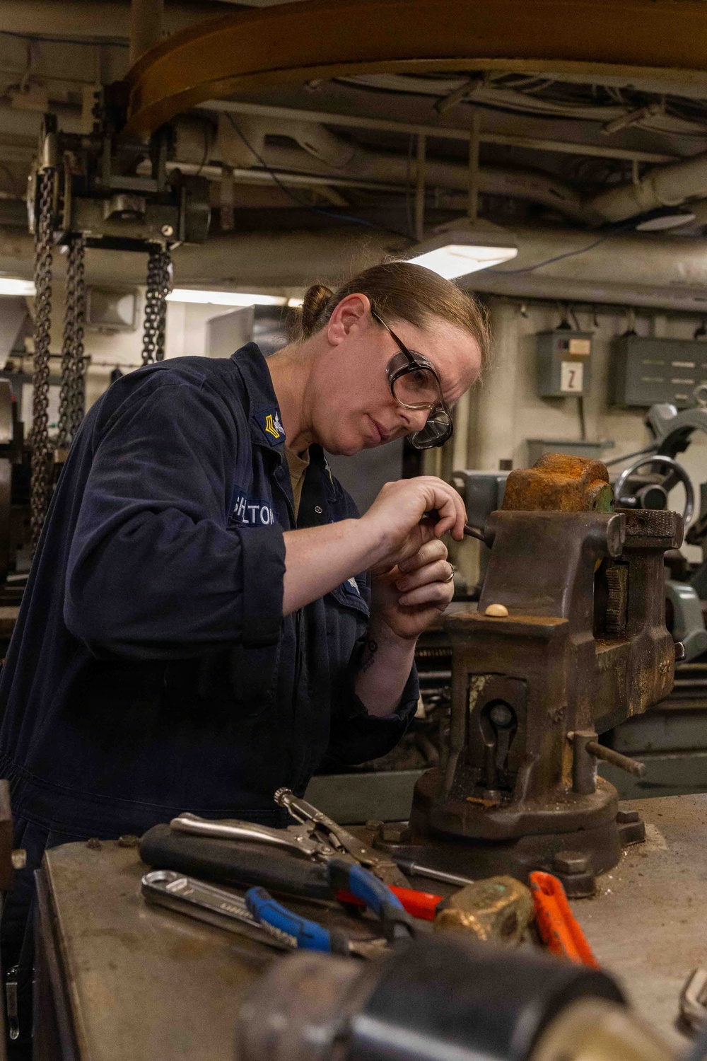 A female military service member wearing a navy blue uniform with a rank insignia patch is working at a workbench in what appears to be a shipboard machine shop or maintenance space. She is wearing safety goggles and is focused on a precision task involving a bench vise. The environment is industrial with overhead chains, piping, and various machinery visible in the background.