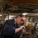 A female military service member wearing a navy blue uniform with a rank insignia patch is working at a workbench in what appears to be a shipboard machine shop or maintenance space. She is wearing safety goggles and is focused on a precision task involving a bench vise. The environment is industrial with overhead chains, piping, and various machinery visible in the background.