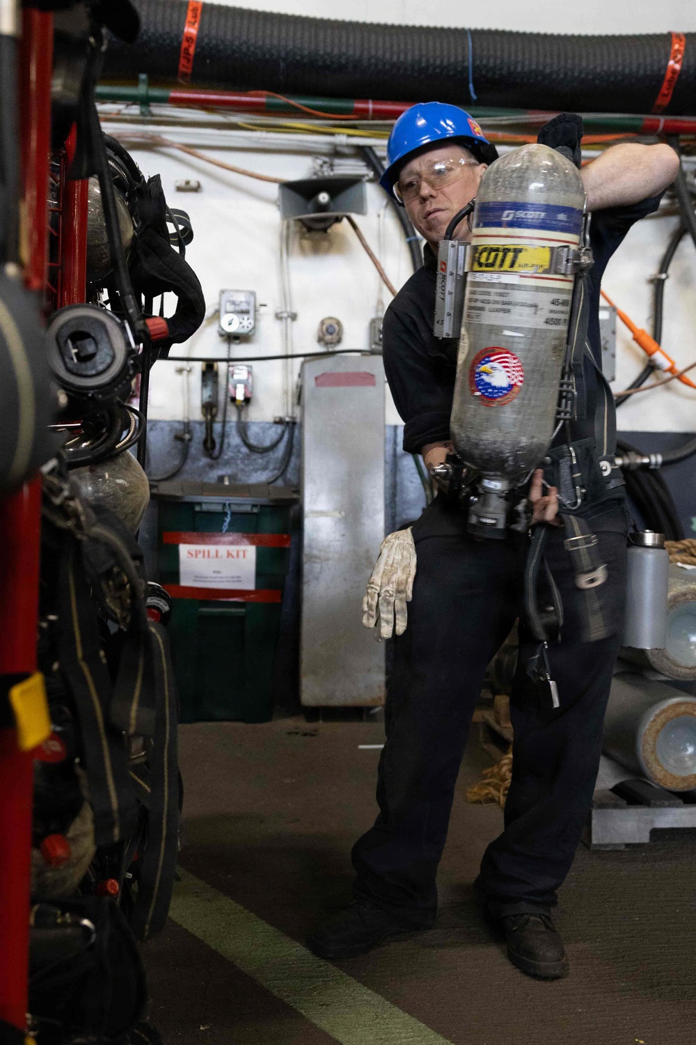 A worker wearing a blue hard hat and safety glasses is handling a Scott brand Self-Contained Breathing Apparatus (SCBA) air tank in what appears to be an industrial or emergency services facility. The person is dressed in dark work clothing with work gloves and is positioned near heavy equipment. The background shows a utility wall with wiring, a spill kit cabinet, and various industrial components.