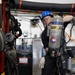 A worker wearing a blue hard hat and safety glasses is handling a Scott brand Self-Contained Breathing Apparatus (SCBA) air tank in what appears to be an industrial or emergency services facility. The person is dressed in dark work clothing with work gloves and is positioned near heavy equipment. The background shows a utility wall with wiring, a spill kit cabinet, and various industrial components.