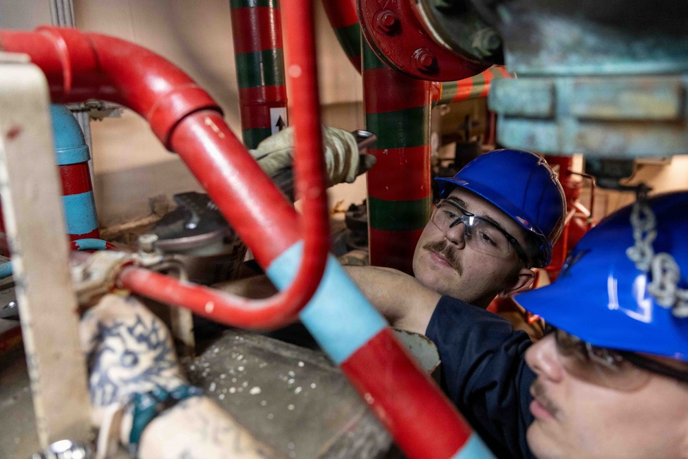 Two workers wearing blue hard hats and safety glasses are inspecting or repairing a complex pipe system in a confined industrial space. The piping includes red and light blue pipes, suggesting a fire suppression or fluid distribution system. One worker has visible tattoos on their arm and is closely examining components between the pipes.