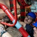 Two workers wearing blue hard hats and safety glasses are inspecting or repairing a complex pipe system in a confined industrial space. The piping includes red and light blue pipes, suggesting a fire suppression or fluid distribution system. One worker has visible tattoos on their arm and is closely examining components between the pipes.