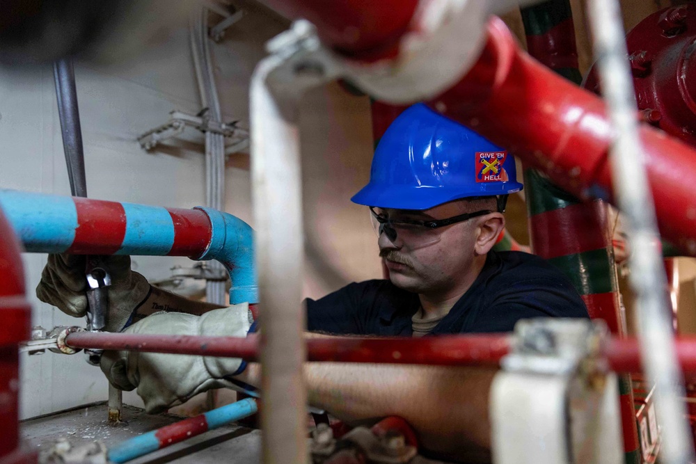 A male worker wearing a blue hard hat with a 'Give 'Em Hell' sticker and safety glasses is performing maintenance or repair work in a confined mechanical space. He is surrounded by a network of red and blue/cyan painted pipes and valves, using a hand tool. The environment appears to be an industrial or naval engineering space.