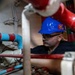 A male worker wearing a blue hard hat with a 'Give 'Em Hell' sticker and safety glasses is performing maintenance or repair work in a confined mechanical space. He is surrounded by a network of red and blue/cyan painted pipes and valves, using a hand tool. The environment appears to be an industrial or naval engineering space.
